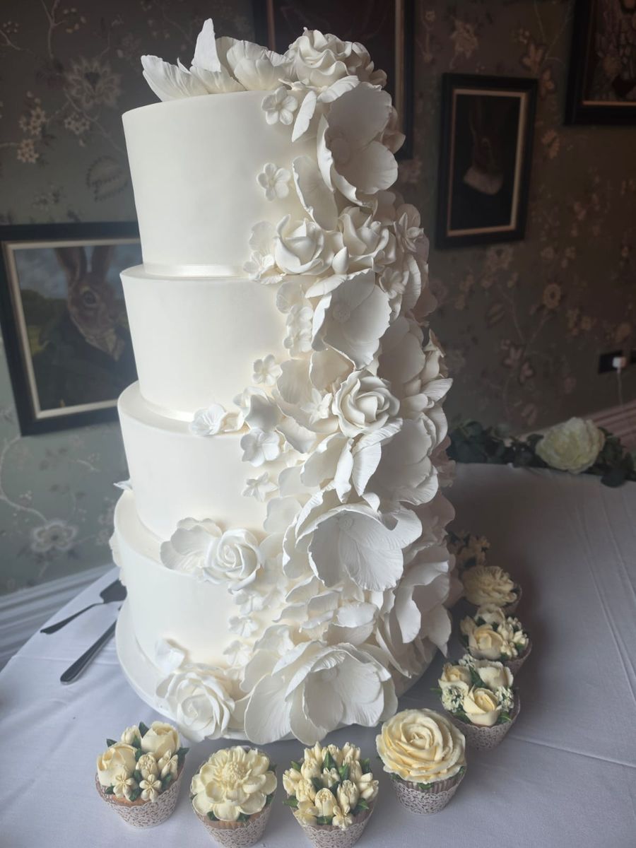 Four tier white wedding cake with a cascade of sugar flowers and matching cupcakes