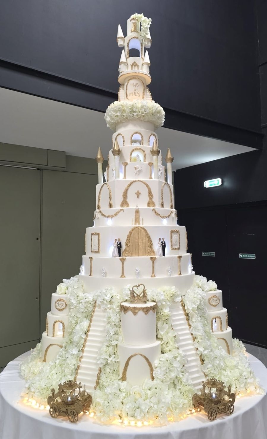 Towering white and gold castle wedding cake with twin staircases, hydrangeas and golden carriages