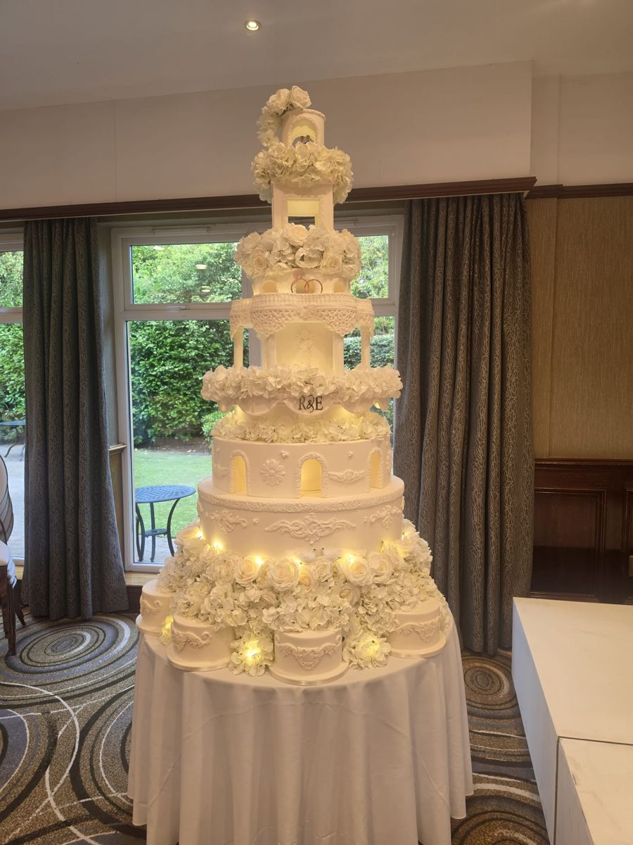 Grand white wedding cake with lit pillared tiers, lace detail and a ring of satellite cakes