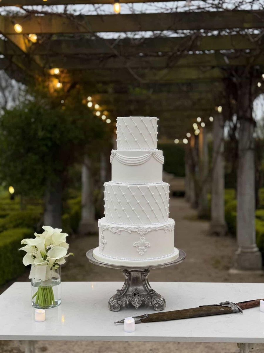 Four tier white wedding cake with quilted pearl detail under a lit garden pergola