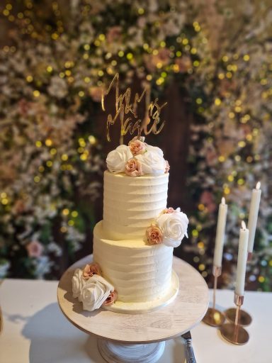 Two-tier wedding cake decorated with flowers and a "Mr & Mrs" cake topper, set against floral backdrop.