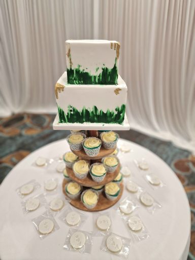 Two-tiered white wedding cake with green decorative accents atop a stand of cupcakes.