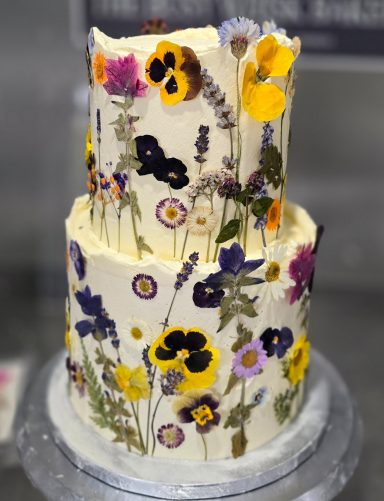 Two-tiered cake decorated with edible flowers and herbs on a white frosting background.
