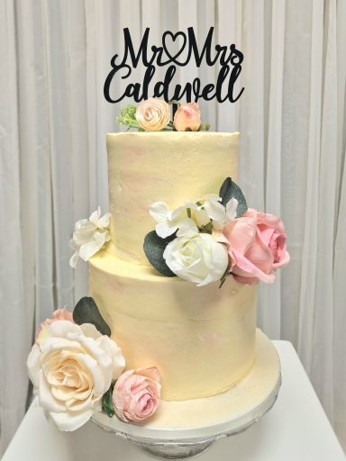 Three-tiered wedding cake with pastel icing, adorned with flowers and a "Mr & Mrs Caldwell" topper.